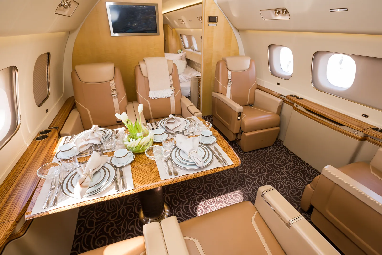 Private jet cabin with a dining table set with china, glassware and white tulips between cream leather seats
